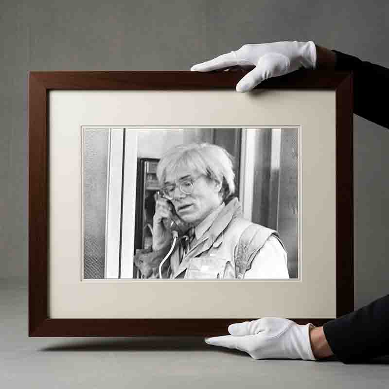 Eine Schwarz-Weiß-Fotografie gerahmt in einem dunklen Holzrahmen mit einem breiten cremefarbenen Passepartout. Das Foto zeigt den Künstler Andy Warhol wie er in einer Telefonzelle telefoniert.