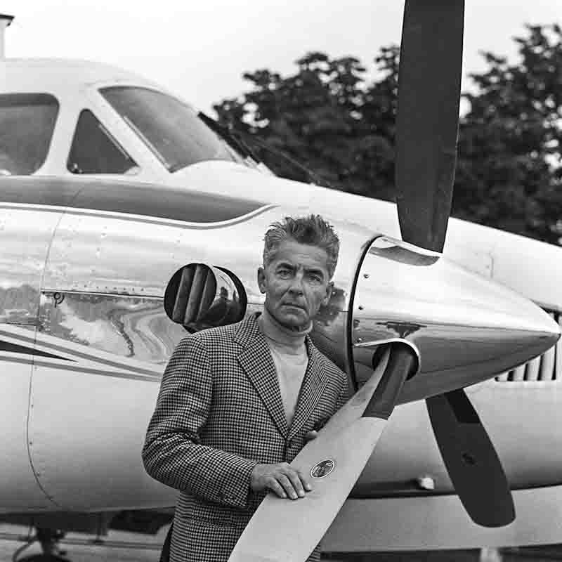 Dirigent Herbert von Karajan im Anzug posiert neben einem Flugzeug, das auf dem Flughafen geparkt ist.