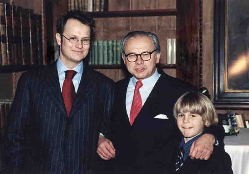 Felix Burda und Hubert Burda in dunklen Anzügen mit Krawatten und ein Jakob Burda stehen lächelnd vor einem Bücherregal.