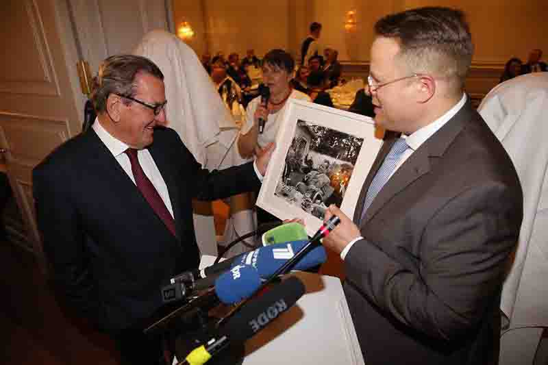 Altkanzler Gerhard Schröder (links) lächelt, während ihm ein gerahmtes Schwarz-Weiß-Foto aus der Sammlung Bock-Schröder überreicht wird.