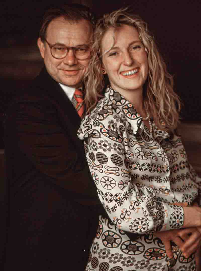 Porträt von Maria Furtwängler und Hubert Burda: Furtwängler lächelt im gemusterten Hemd vorne, Burda steht im dunklen Anzug mit Krawatte hinter ihr und umarmt sie.