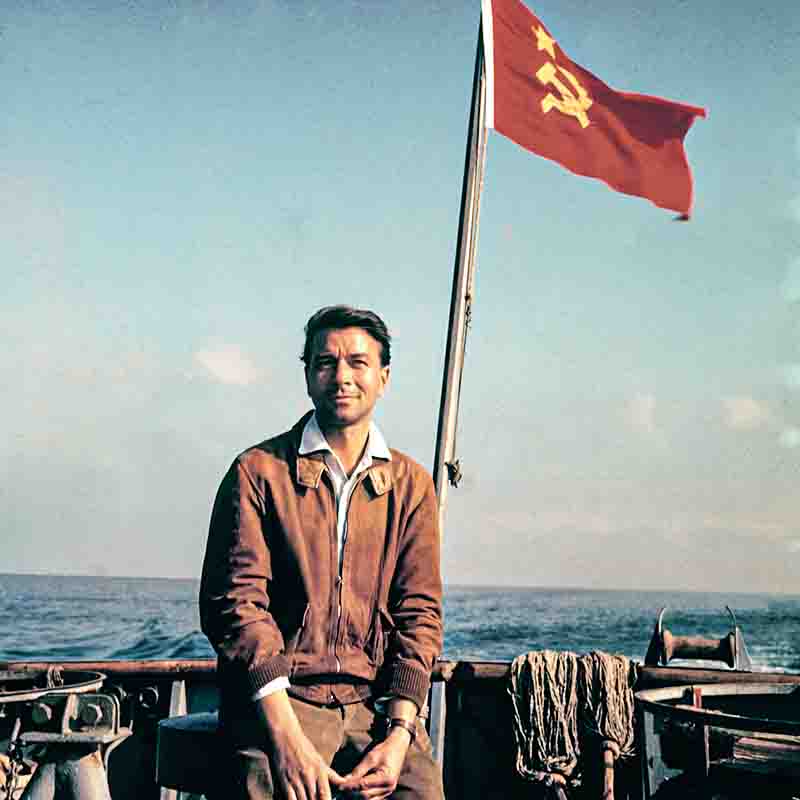 Der deutsche Fotojournalist Peter Bock-Schroeder sitzt auf einem Schiff auf dem Meer, hinter ihm weht die Flagge der Sowjetunion am Mast.