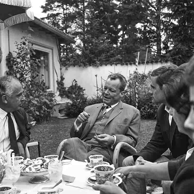 Ein Schwarz-Weiß-Foto zeigt Willy Brandt, den Bundeskanzler der Bundesrepublik Deutschland (1969 bis 1974), der im Freien an einem Tisch mit einer kleinen Gruppe von Menschen sitzt, anscheinend während eines gesellschaftlichen oder informellen Treffens.